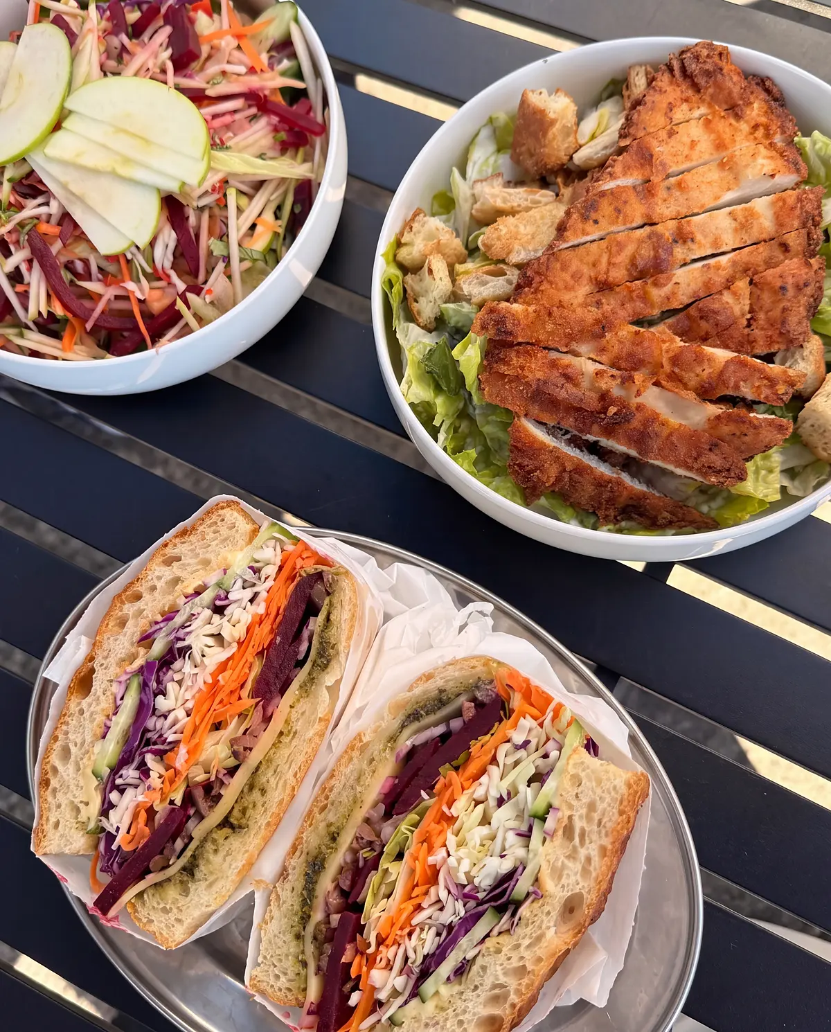 Two salad focaccias cut open on a steel tray, with a harvest bowl and a caesar bowl, from above on a black table